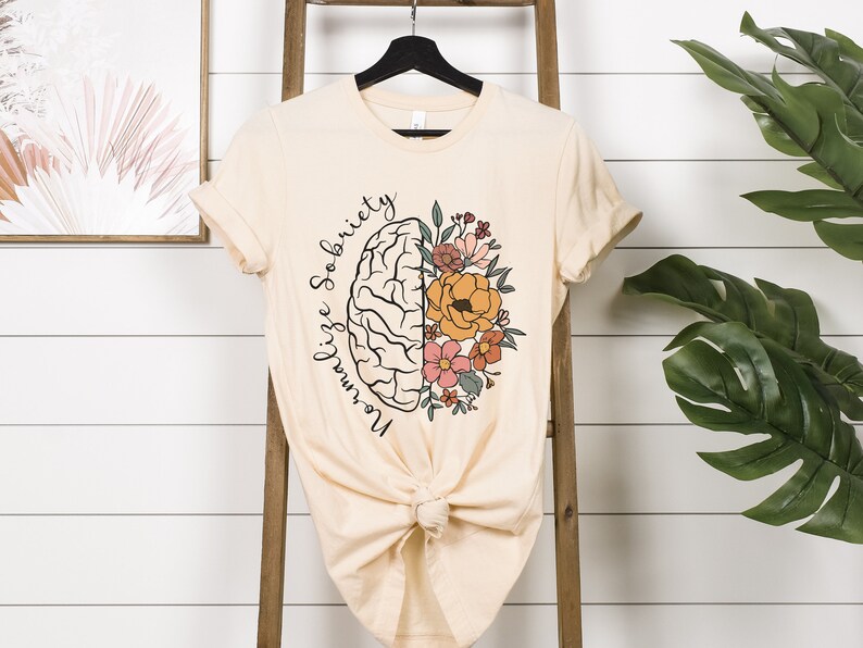 May include: Cream-colored t-shirt featuring a graphic of a brain split in half, with one side a brain and the other side flowers. The text "Normalize Sobriety" is written in a script font around the brain. The shirt is knotted at the bottom.