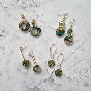 Puede incluir: Un surtido de pendientes dorados con detalles de piedra moteada verde, marrón y dorada. Los pendientes varían en diseño, incluyendo aros, círculos colgantes y estilos lineales. Los pendientes se exhiben sobre una superficie con diseño de mármol.
