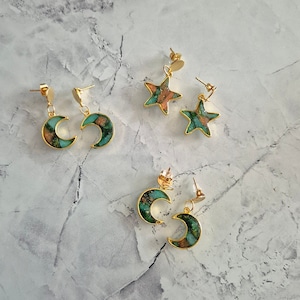 Op de afbeelding: Een set van vier gouden oorbellen met groene en bruine harsinlegsels. Twee oorbellen zijn halvemaanvormig en twee zijn stervormig.