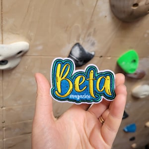 May include: A colorful embroidered patch with the word "Beta" in yellow script outlined in blue, with the word "magazine" below. The patch has a white border and is held against a climbing wall.