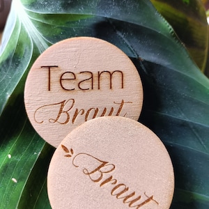 Könnte beinhalten: Zwei runde Holzschilder mit dem Text "Team Braut" und "Braut" in Kursivschrift. Die Schilder liegen auf einem grünen Blattwerk.