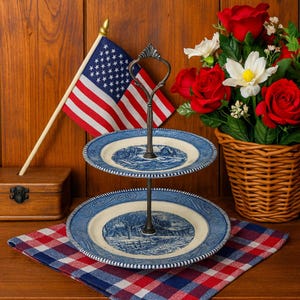 May include: A two-tiered serving stand with blue and white plates, featuring a landscape design. An American flag and a basket of red roses and white flowers are in the background. The stand is on a red and blue checkered cloth.