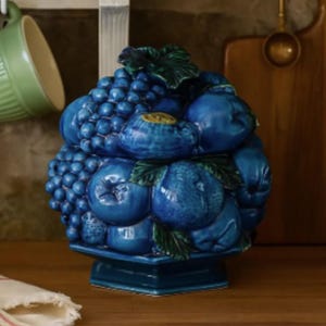 May include: A blue ceramic fruit-shaped container with a lid, featuring grapes, blueberries, and other fruits. The container is on a square base and has green leaves. The background includes a wooden cutting board and a green mug.