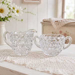 May include: A clear glass creamer pitcher and a matching sugar bowl with two handles. Both pieces feature a faceted, geometric diamond-cut pattern across their exterior surfaces, reflecting light through the transparent glass material.