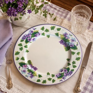 May include: A round white ceramic dinner plate featuring a decorative border of purple violet flowers and green ivy leaves. The plate has a thin purple rim and a smooth, glossy finish, centered on a table setting with a fork and a knife.
