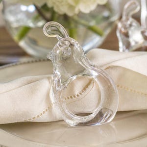 May include: A clear glass bunny-shaped napkin ring. The ring is on a white napkin, with a blurred background of flowers in a vase. The bunny has a detailed head and ears, and the ring is a circular shape.