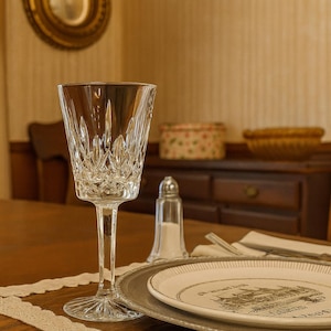 May include: A clear crystal wine glass with a detailed cut pattern stands on a wooden table. A silver-rimmed plate with a decorative design, a salt shaker, and silverware are also on the table. The background features a wooden cabinet and a decorative mirror.