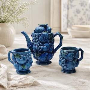 May include: A glossy cobalt blue ceramic tea set consisting of a teapot and two matching mugs. Each piece features a three-dimensional relief design of various fruits, including grapes, apples, and pears, with small green leaves accenting the fruit.