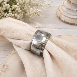 May include: A wide, polished silver-toned metal napkin ring with a smooth, curved surface and subtle raised detailing along the edges. The ring is wrapped around a folded, light beige linen napkin, showcasing a reflective and metallic finish.