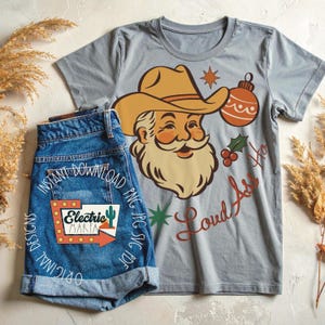 May include: A gray t-shirt with a Santa Claus graphic wearing a cowboy hat, a Christmas ornament, and the text "Loud Ass Yo". Paired with denim shorts featuring a "Electric Marfa" design.