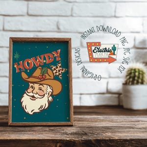 May include: A framed print featuring a cartoon Santa Claus wearing a cowboy hat with the word "HOWDY" in orange and red. The background is teal with a star and a cactus logo. The print is in a wooden frame.