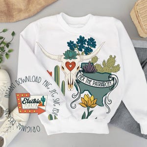 May include: White sweatshirt featuring a graphic of a bull skull with a heart, cacti, and the text "Hex the Patriarchy." The design includes blue, green, and yellow accents. The sweatshirt has long sleeves and a crew neck.