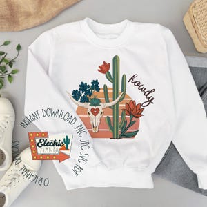 May include: White sweatshirt featuring a desert-themed graphic. The design includes a steer skull with a heart, cactus, and the word "howdy." The design is in shades of orange, green, and blue. The sweatshirt also has the text "Instant Download" and "Electric Marfa."
