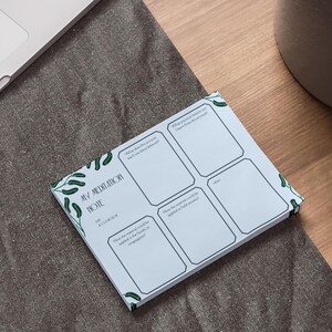 May include: A light blue meditation note pad with a green leaf design. The notepad has six rectangular sections for writing. The text "MY MEDITATION NOTE" is printed on the pad. The notepad is on a wooden desk next to a laptop and a plant.