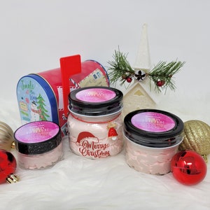 May include: Three glass jars filled with pink body butter, labeled "Body Butter". The jars have black lids and are decorated with a Christmas theme. The middle jar has a red Santa hat and the text "Oh Merry Christmas".