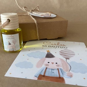 May include: A small glass bottle with a wooden lid and a yellow liquid inside. The bottle has a label that says "Carlos Mi Bautizo". The bottle is tied with twine to a small wooden bead. There is a card with a cartoon bunny holding a balloon that says "Carlos Mi Bautizo".