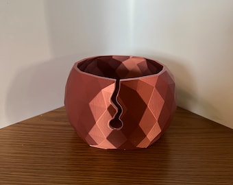 May include: A copper-colored, geometric-patterned planter with a unique, curved opening. The spherical shape is divided into diamond-shaped facets, creating a modern, textured appearance. The planter sits on a wooden surface.