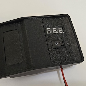 以下が含まれることがあります： 「8.8.8」と表示されたデジタルディスプレイとオン/オフスイッチを備えた黒い電子デバイス。赤いワイヤーが下から出ています。デバイスは長方形で、左側にくぼんだ領域があります。