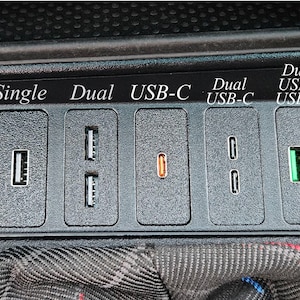May include: A black car console featuring various USB ports. The console includes single and dual USB ports, along with USB-C ports, labelled "Single", "Dual", and "USB-C". There is also a circular opening.
