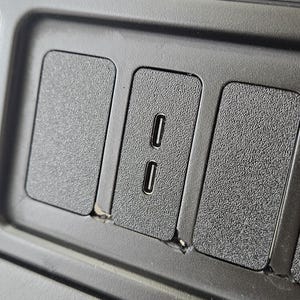 May include: Close-up of a black plastic car console with textured surfaces. Features include two USB-C ports and several rectangular, textured panels. The console is likely part of a vehicle's interior.