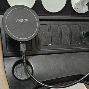 May include: A black VIKEFON wireless charging pad is connected to a car's console. The pad is round with a white border and the brand name in white. A black USB cable plugs into the console's USB port.