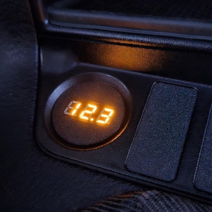 Puede incluir: Primer plano del salpicadero de un coche con una pantalla digital iluminada que muestra "82.3". La pantalla es naranja y está situada en una carcasa circular negra. Adyacentes a la pantalla hay tres paneles rectangulares texturizados.