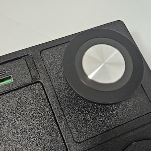 May include: Black textured surface with a circular silver and black control knob. A USB-C port with a green interior is visible. The item appears to be a desk accessory or a component of a larger device.