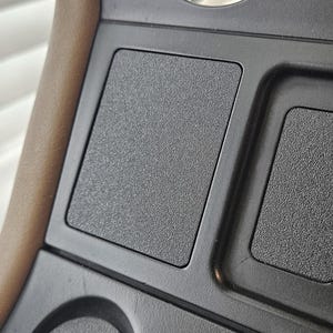 May include: Close-up of a black textured car console with rectangular compartments. The console has a matte finish and is set against a blurred background. The design suggests an automotive interior.