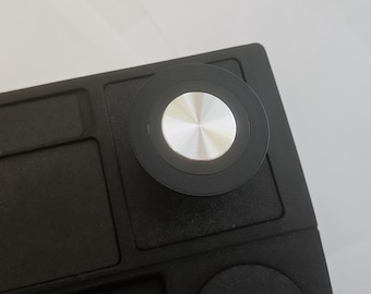 May include: Close-up of a black rectangular object with a circular, silver-colored button. The button is centered on the top right corner of the object. The object has a textured surface and a recessed area.