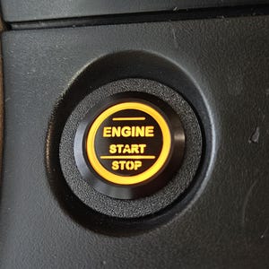 Op de afbeelding: Een close-up van de start/stop-knop van een auto. De knop is zwart met een geel verlichte ring en tekst. De tekst luidt "ENGINE START STOP". De knop is geplaatst in een zwart paneel.
