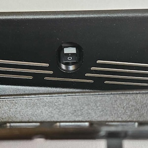 May include: A black rectangular component with a recessed area and a small on/off switch. The component has several horizontal silver bars and a textured surface.