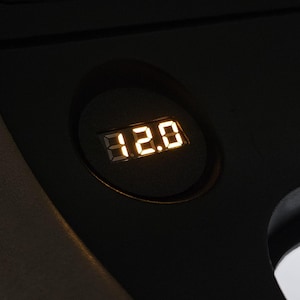 Puede incluir: Primer plano de una pantalla digital que muestra el número 12,0 en color amarillo-naranja brillante. La pantalla está en una carcasa circular negra, probablemente parte del salpicadero o la consola de un vehículo.