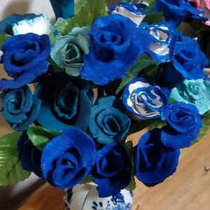 May include: A bouquet of blue and teal roses in a white and blue ceramic vase. The roses are arranged in a heart shape.