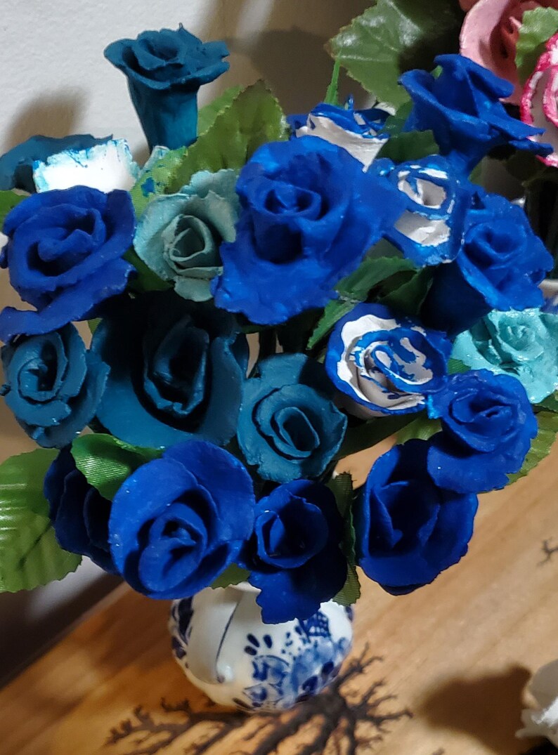 May include: A bouquet of blue and teal roses in a white and blue ceramic vase. The roses are arranged in a heart shape.