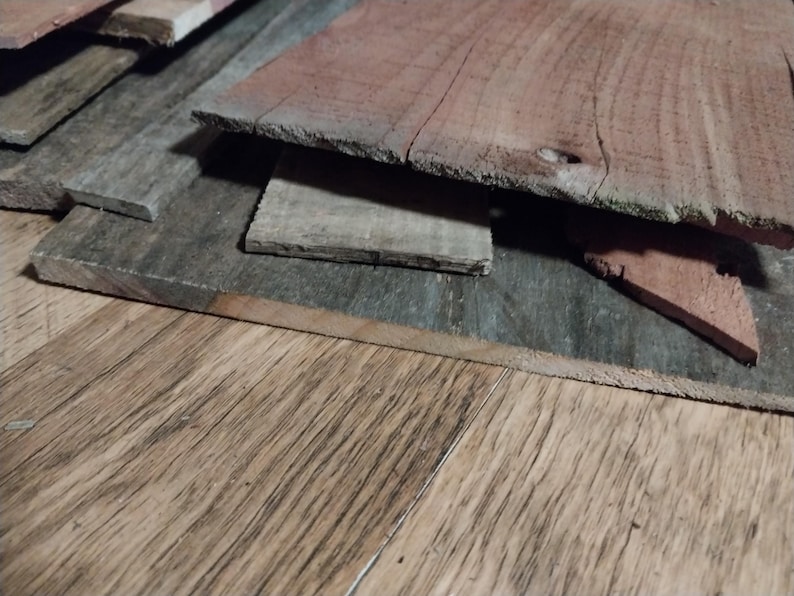May include: A collection of weathered wooden planks in various shades of gray and reddish-brown. The planks are stacked, revealing their rough, textured surfaces and natural grain patterns. The image suggests a rustic or reclaimed wood theme.