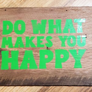 May include: A rectangular wooden sign with the words "DO WHAT MAKES YOU HAPPY" in bright green lettering. The sign is made from weathered wood, showing the natural grain and knots. The sign has a natural wood finish.
