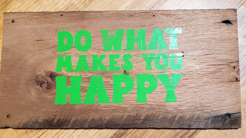 May include: A rectangular wooden sign with the words "DO WHAT MAKES YOU HAPPY" in bright green lettering. The sign is made from weathered wood, showing the natural grain and knots. The sign has a natural wood finish.