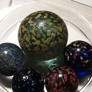 Op de afbeelding: Vijf glazen knikkers met kleurrijke, gevlekte patronen. De knikkers zijn groen, blauw, zwart en rood.