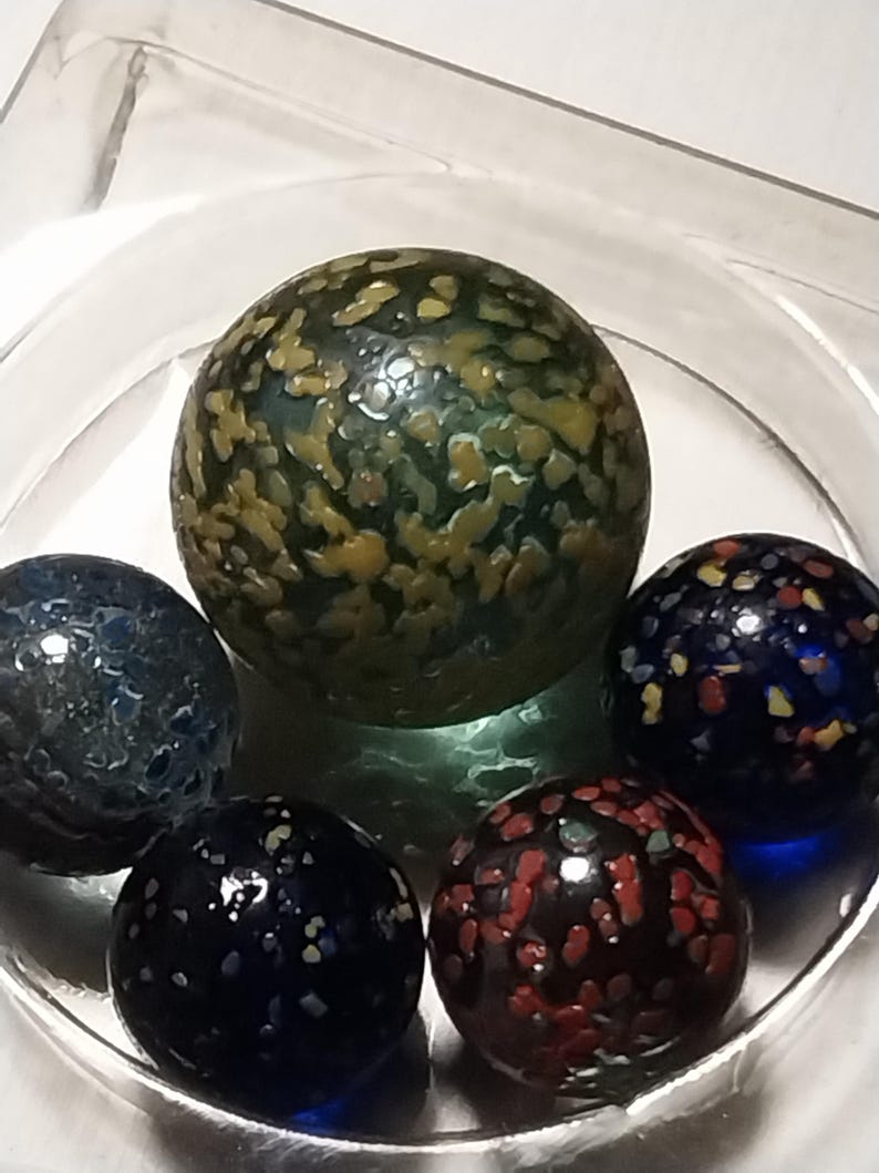 Op de afbeelding: Vijf glazen knikkers met kleurrijke, gevlekte patronen. De knikkers zijn groen, blauw, zwart en rood.