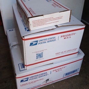 May include: Three white cardboard boxes stacked on top of each other. The top box is a Priority Mail box with the text "PRIORITY MAIL" in red letters. The middle box is also a Priority Mail box with the text "PRIORITY MAIL" in red letters. The bottom box is a Large Flat Rate Box with the text "LARGE FLAT RATE BOX" in black letters. All three boxes have the United States Postal Service logo on them.