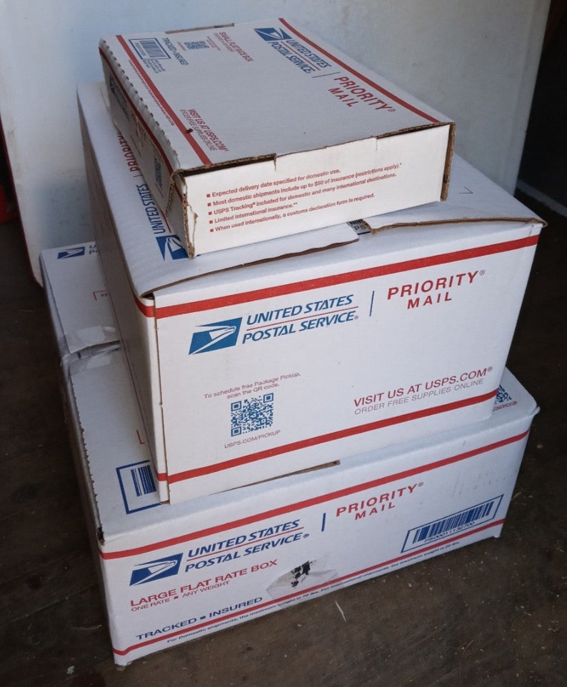 May include: Three white cardboard boxes stacked on top of each other. The top box is a Priority Mail box with the text "PRIORITY MAIL" in red letters. The middle box is also a Priority Mail box with the text "PRIORITY MAIL" in red letters. The bottom box is a Large Flat Rate Box with the text "LARGE FLAT RATE BOX" in black letters. All three boxes have the United States Postal Service logo on them.