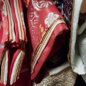 May include: Close-up of ornate Japanese kimono fabric. The red fabric features white floral patterns and silver accents. Gold and white trim borders the sleeves and edges. The image showcases the intricate details and textures of the traditional garment.