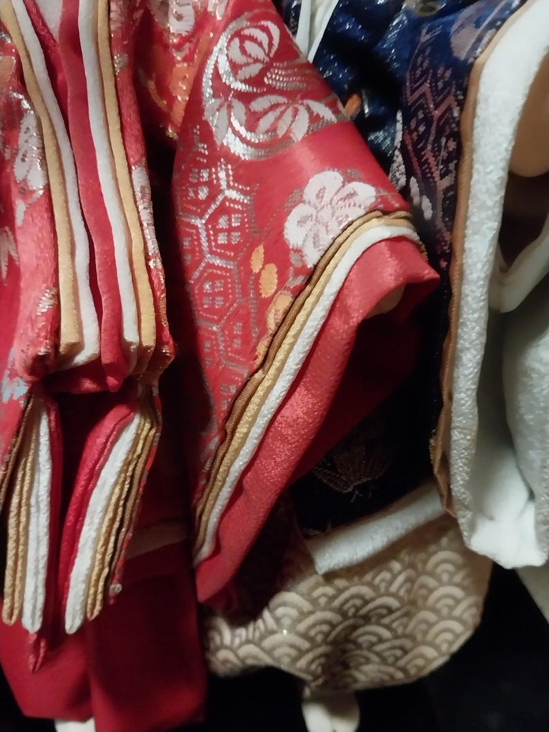May include: Close-up of ornate Japanese kimono fabric. The red fabric features white floral patterns and silver accents. Gold and white trim borders the sleeves and edges. The image showcases the intricate details and textures of the traditional garment.