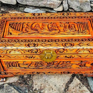 May include: A rectangular wooden chest with intricate carvings and a brass clasp. The chest is stained in a warm orange hue, with detailed relief carvings of sailing ships and floral patterns. The chest rests on small, square feet.