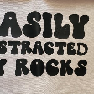 May include: A cream-colored fabric banner with the phrase "EASILY DISTRACTED BY ROCKS" printed in large, black, bubble letters. The banner is partially visible, with a wooden surface in the background.