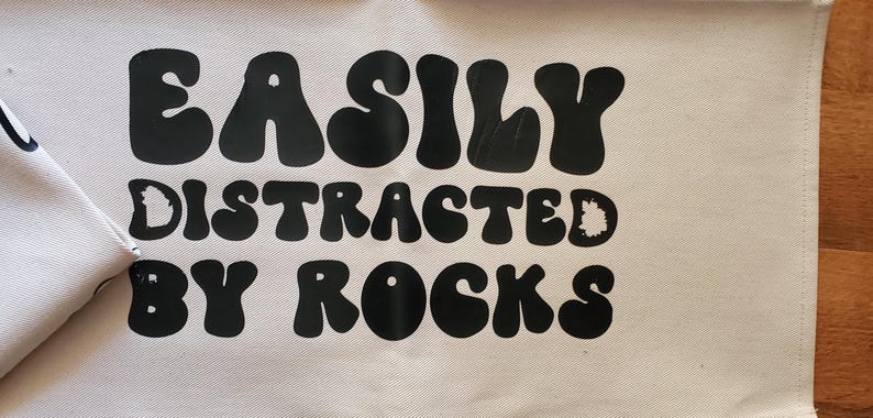 May include: A cream-colored fabric banner with the phrase "EASILY DISTRACTED BY ROCKS" printed in large, black, bubble letters. The banner is partially visible, with a wooden surface in the background.
