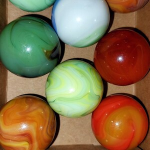 May include: A collection of colorful glass marbles in a cardboard box. The marbles feature swirls of green, orange, red, and white. The marbles are round and have a glossy finish, showcasing their vibrant colors and patterns.