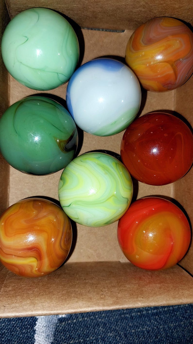 May include: A collection of colorful glass marbles in a cardboard box. The marbles feature swirls of green, orange, red, and white. The marbles are round and have a glossy finish, showcasing their vibrant colors and patterns.