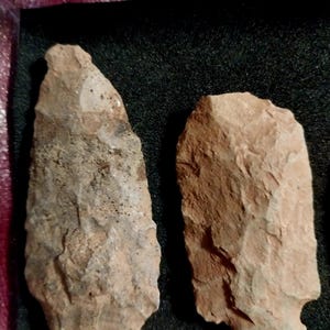 May include: Two brown stone arrowheads, one with a more pointed tip and the other with a more rounded tip, are displayed on a black background.