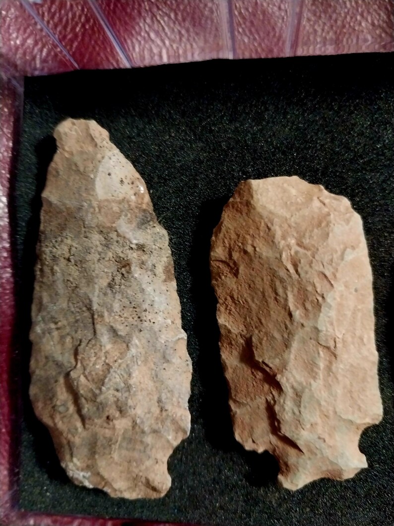 May include: Two brown stone arrowheads, one with a more pointed tip and the other with a more rounded tip, are displayed on a black background.